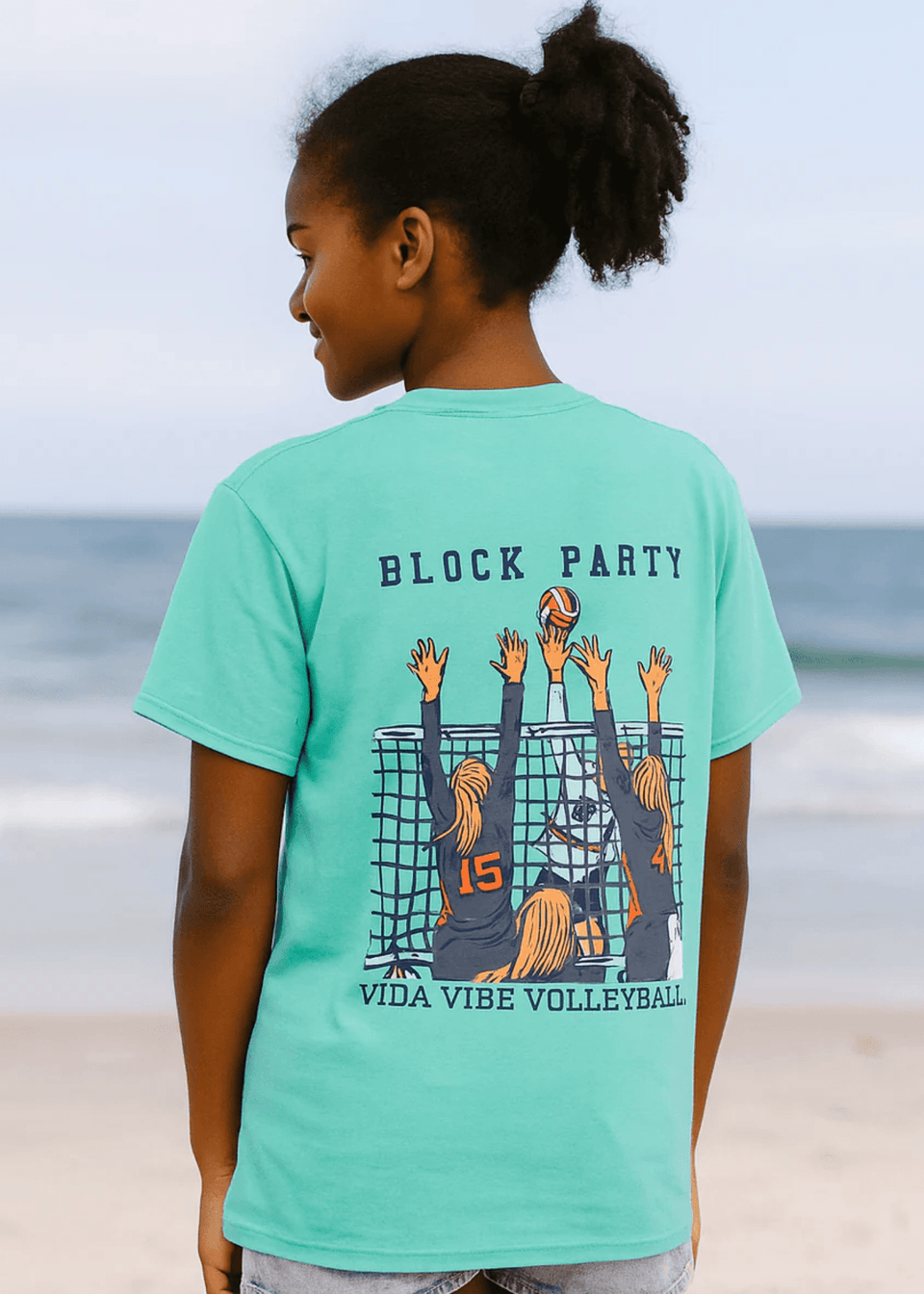 Child wearing a teal t-shirt with 'Block Party' and volleyball design on a beach.